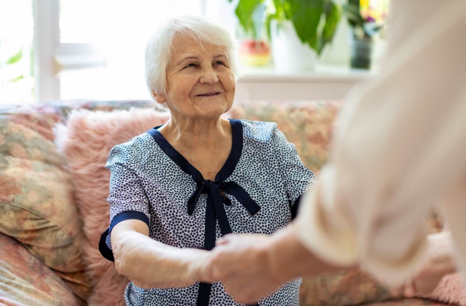 An older adult smiles at a caregiver while grasping their hands and preparing to stand up from a couch