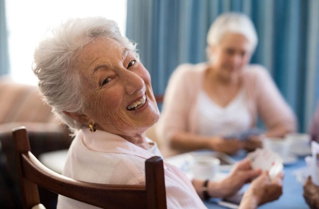 An older adult turns to grin at the camera while playing cards with other residents in the common lounge of an assisted living community