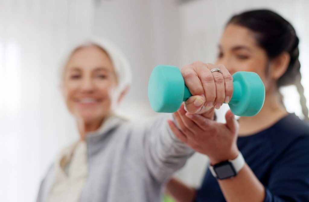 A physical trainer helps a smiling older adult lift a small blue weight safely with proper form