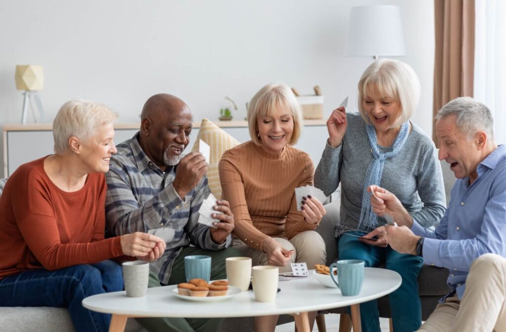 5 older adults laugh and play cards over tea and muffins together in independent living