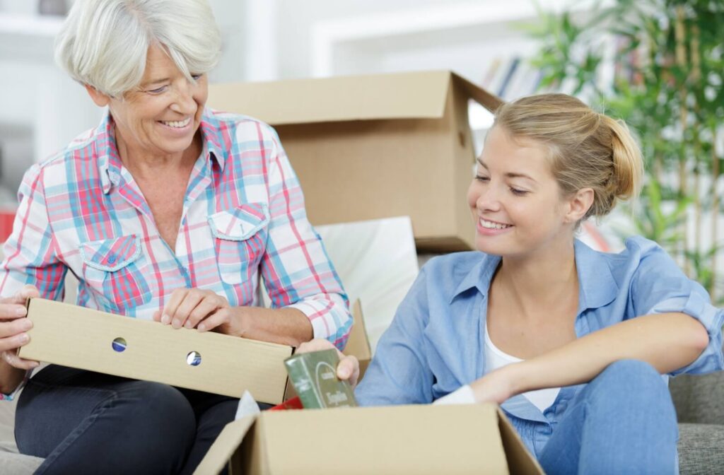 An adult child sits beside their older parent and smiles while unpacking a box of books and sitting on the floor in an independent living apartment