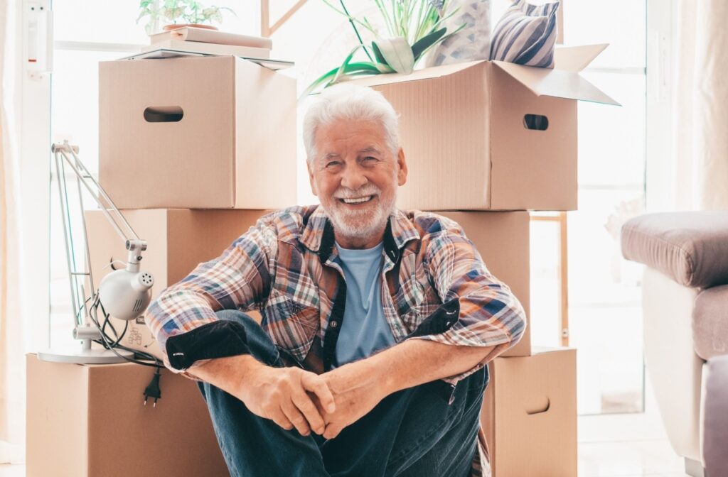 A senior smiles as they sit in front of stacks of boxes packed with their belongings, excited to move into their new senior living community.

