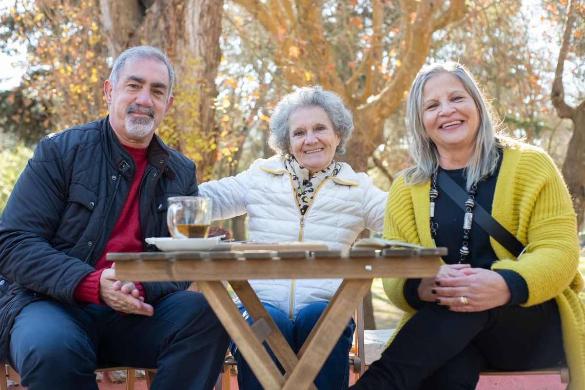 Three older adults sit smiling around a small wooden table outdoors among autumn trees, with a glass of tea.