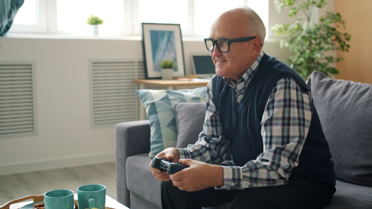 An older adult in glasses and a knit vest sits on a grey sofa holding a game controller, smiling, with mugs on a table in the foreground.
